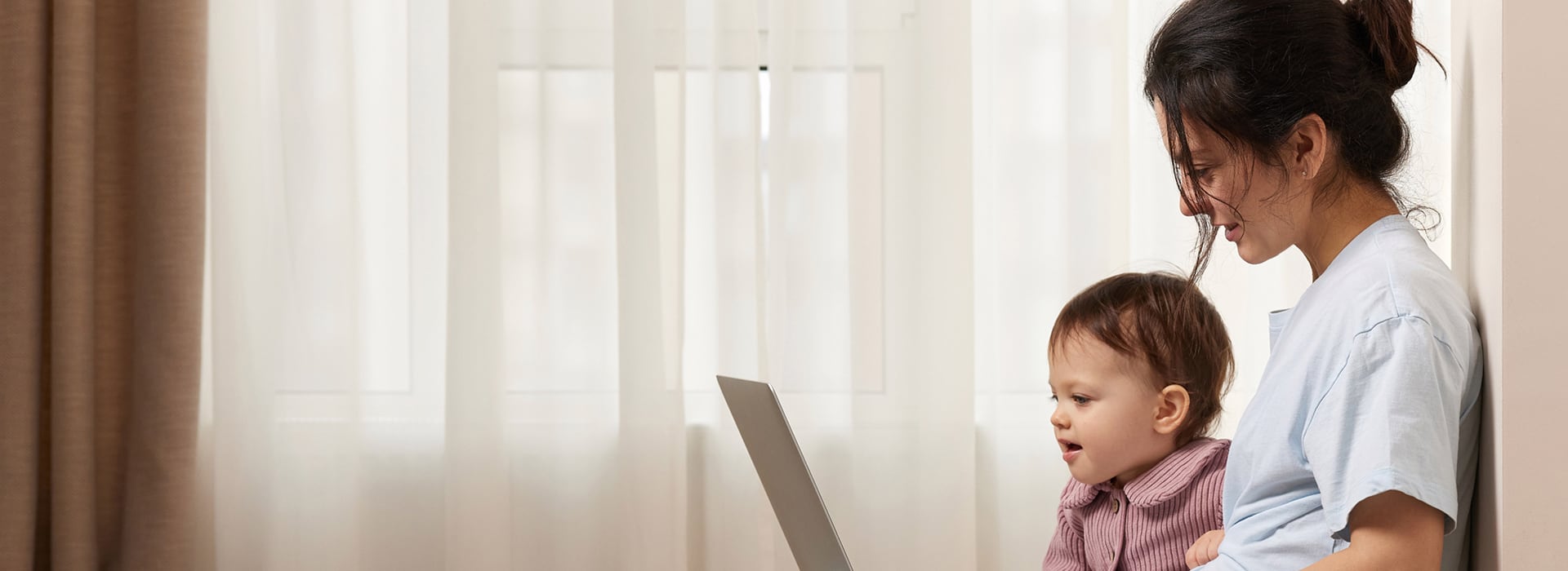 A woman sits on a wooden floor next to an infant with a laptop on her lap.